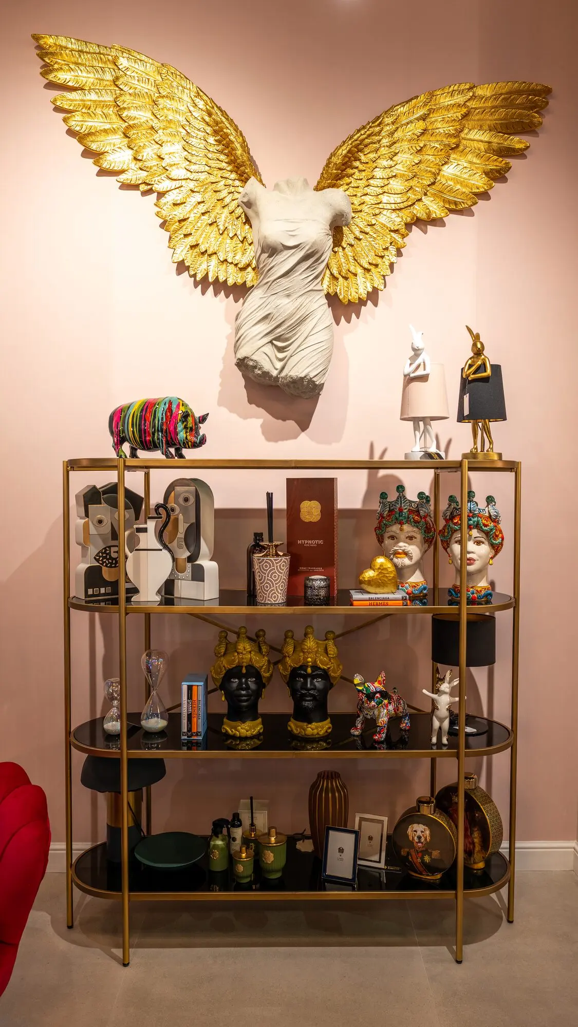 Busto bianco con grandi ali dorate sopra una scaffalatura di ceramiche e oggetti decorativi, su una parete rosa.