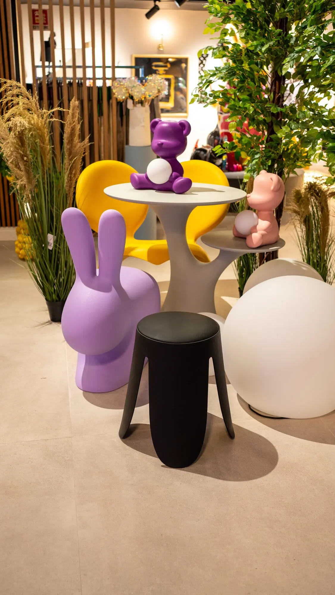 Sedute a forma di coniglio lilla e sgabello nero accanto a un tavolo con un orsetto viola e sedie gialle.