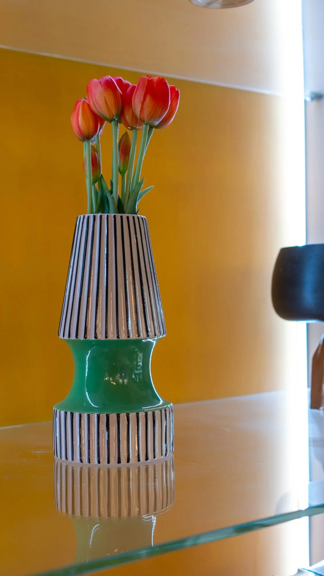 Vaso in ceramica a righe bianche e nere con base verde e un mazzo di tulipani rossi, su un ripiano di vetro.